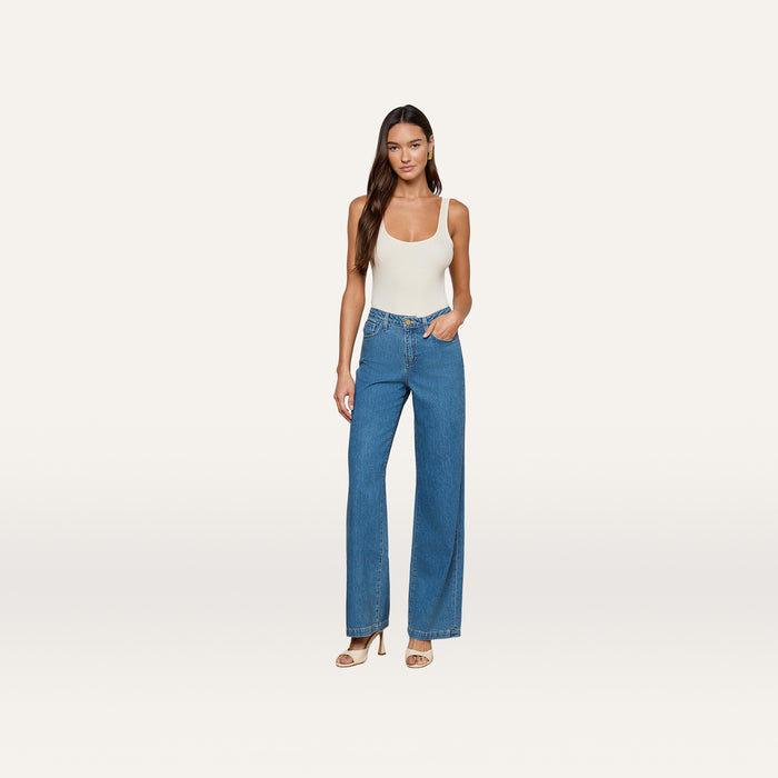A woman stands against a plain background, wearing a white tank top, the Scottie Wide-Leg Jean in medium wash blue high-waisted all-cotton denim, and beige heeled sandals. She has long hair and is posing with one hand in her pocket.