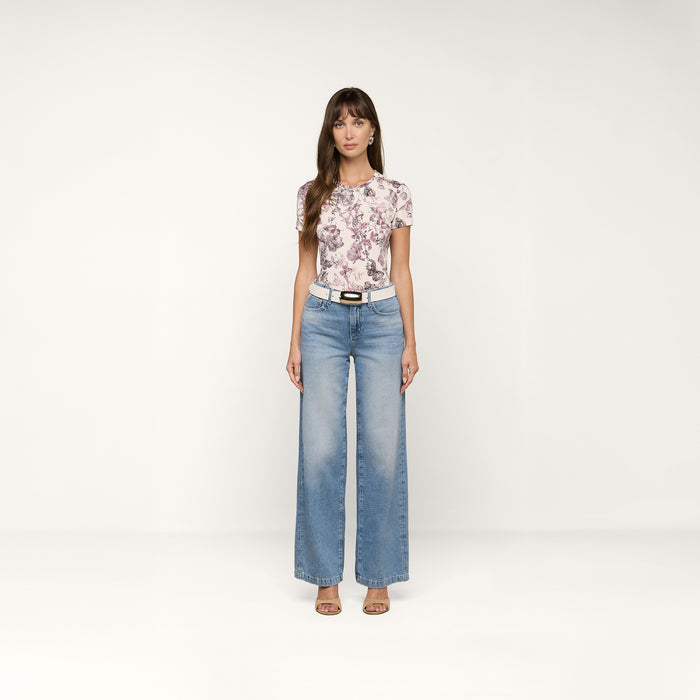 A woman stands facing forward wearing a floral short-sleeve top, a white belt, sandals, and the Scottie Wide-Leg Jean in light blue high-rise all-cotton denim against a plain white background.
