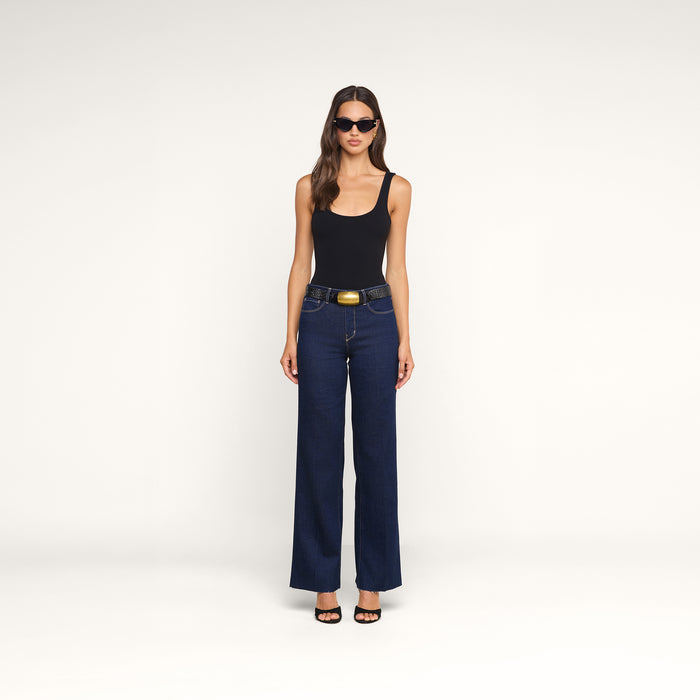 A woman wearing sunglasses, a black tank top, the Scottie Wide-Leg Jean in blue high-rise premium stretch denim with a large belt buckle, and black heels stands against a plain white background.
