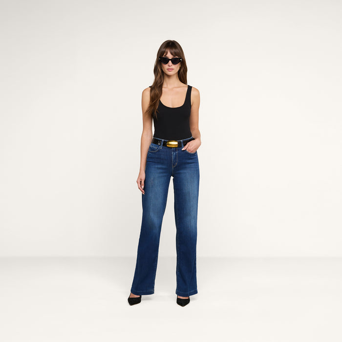 A woman stands against a plain white background, posing with one hand in her pocket, wearing a black tank top, the Scottie Wide-Leg Jean featuring stretch denim and a contoured waistband, paired with black heels and sunglasses.