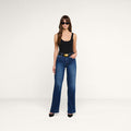 A woman stands against a plain white background, posing with one hand in her pocket, wearing a black tank top, the Scottie Wide-Leg Jean featuring stretch denim and a contoured waistband, paired with black heels and sunglasses.