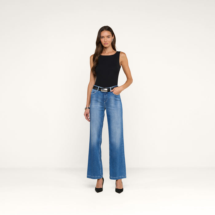 A woman wearing a sleeveless black top, the Scottie Wide-Leg Jean in high-rise blue stretch denim, a black belt, and black heels stands against a plain white background.