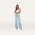 A woman stands against a plain white background, smiling with her arms crossed, wearing a white tank top, beige heels, and the Scottie Wide-Leg Jean in light blue stretch denim featuring a contoured waistband.