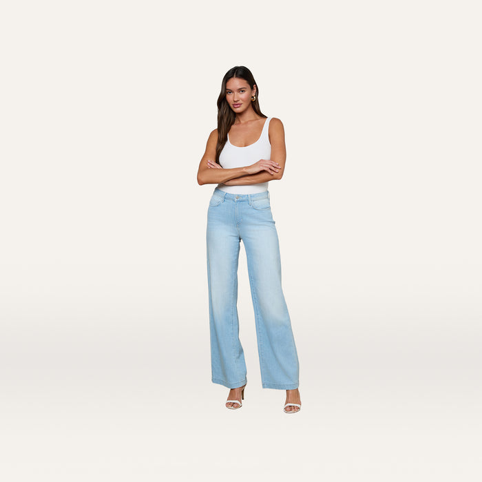 A woman stands against a plain white background, smiling with her arms crossed, wearing a white tank top, beige heels, and the Scottie Wide-Leg Jean in light blue stretch denim featuring a contoured waistband.