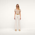 A woman wearing beige sunglasses, a short-sleeve beige top, the Scottie Wide-Leg Jean in cream with a tan belt, and beige heels stands against a plain light background.