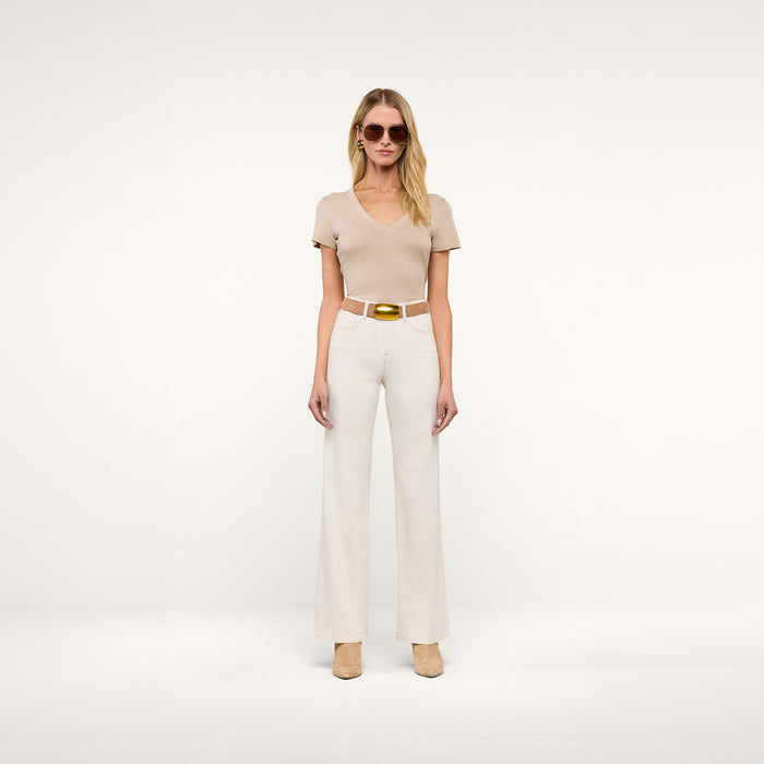 A woman wearing beige sunglasses, a short-sleeve beige top, the Scottie Wide-Leg Jean in cream with a tan belt, and beige heels stands against a plain light background.