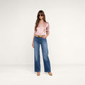 A woman stands confidently against a plain white background, wearing a pink satin blouse, the Scottie Wide-Leg Jean in high-rise blue stretch denim, tan heels, and sunglasses.
