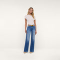 A woman stands against a plain white background wearing a fitted white short-sleeve top, the Scottie Wide-Leg Jean in high-rise blue with a raw hem, a brown belt, and beige heels. She has long blonde hair and a neutral expression.