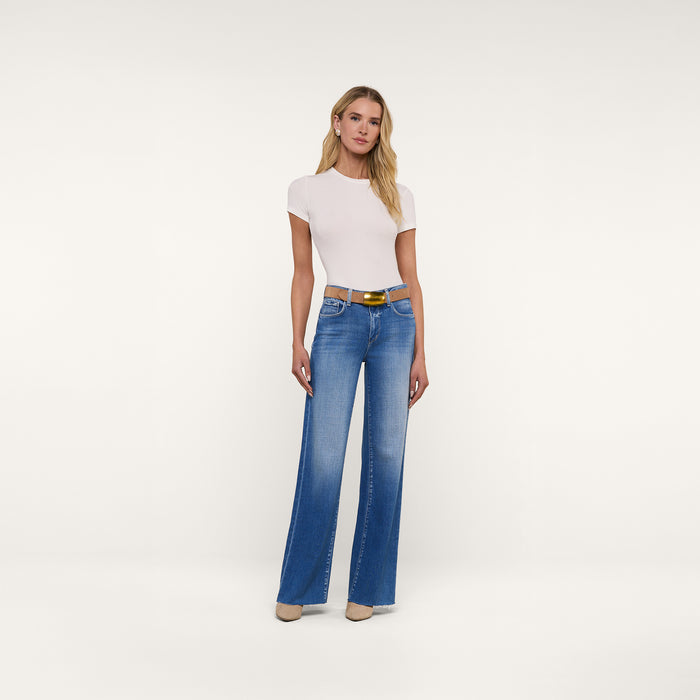A woman stands against a plain white background wearing a fitted white short-sleeve top, the Scottie Wide-Leg Jean in high-rise blue with a raw hem, a brown belt, and beige heels. She has long blonde hair and a neutral expression.
