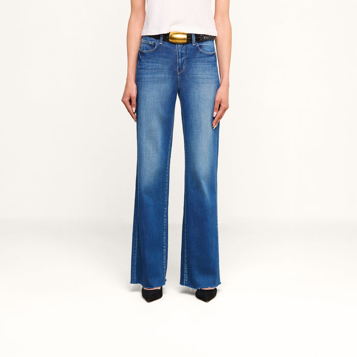 Wearing the Scottie Wide-Leg Jean in vintage-wash denim, a white top, a black belt with a gold buckle, and black pointed shoes, the person stands against a plain white background.
