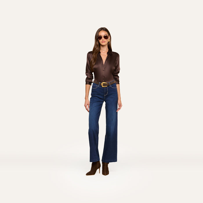 Woman wearing sunglasses, a dark brown satin button-up shirt, the Scottie Wide-Leg Jean in super stretch denim, a brown belt, and brown ankle boots stands against a plain white background.