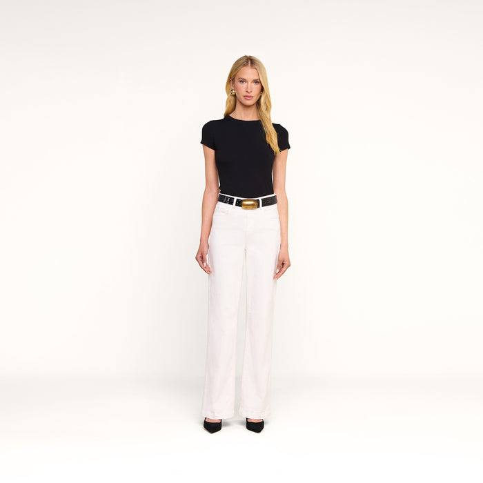 A woman stands against a plain white background, wearing a black short-sleeve top, the Scottie Wide-Leg Jean in white with a high-rise waistband, a black belt with a gold buckle, and black pointed shoes.
