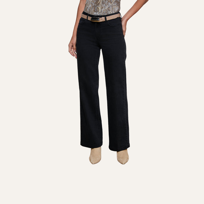 Wearing the Scottie Wide-Leg Jean featuring a contoured waistband, a person styles the black jeans with a beige belt and tan heeled shoes, striking a pose with one hand on their hip against a plain, light-colored background.
