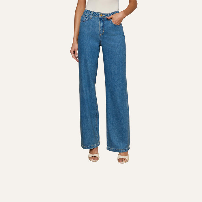 A person shown from the shoulders down against a plain light background wears Scottie Wide-Leg Jean in medium wash, crafted from all-cotton denim, paired with open-toe beige heels.