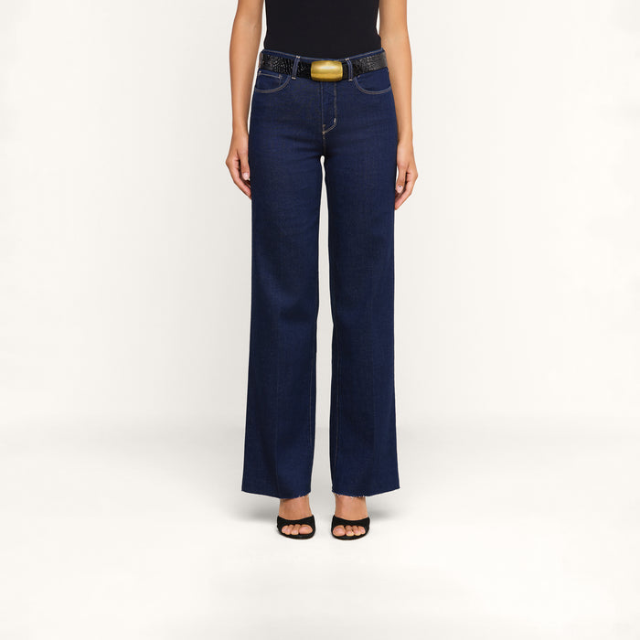 A person wearing the Scottie Wide-Leg Jean in dark blue premium stretch denim, styled with a black belt featuring a large buckle, a black top, and black open-toe heels stands against a plain white background.