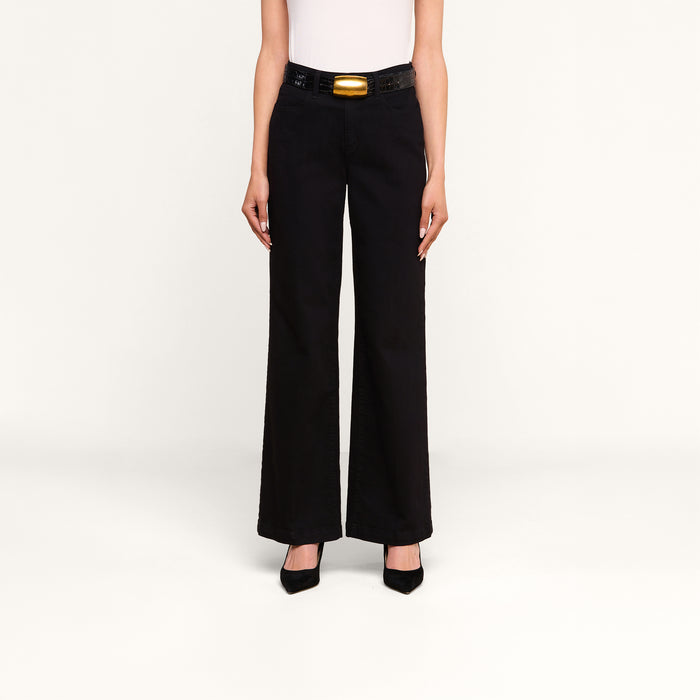 A person stands against a plain background, wearing the Scottie Wide-Leg Jean—a pair of black high-rise jeans crafted from premium stretch denim—paired with a black belt featuring a gold buckle, a white top, and black pointed-toe heels. Their arms are relaxed at their sides.