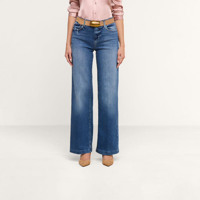 A person wearing the Scottie Wide-Leg Jean in high-rise blue stretch denim, paired with a light pink satin blouse, a tan belt, and tan pointed-toe heels stands against a plain white background.