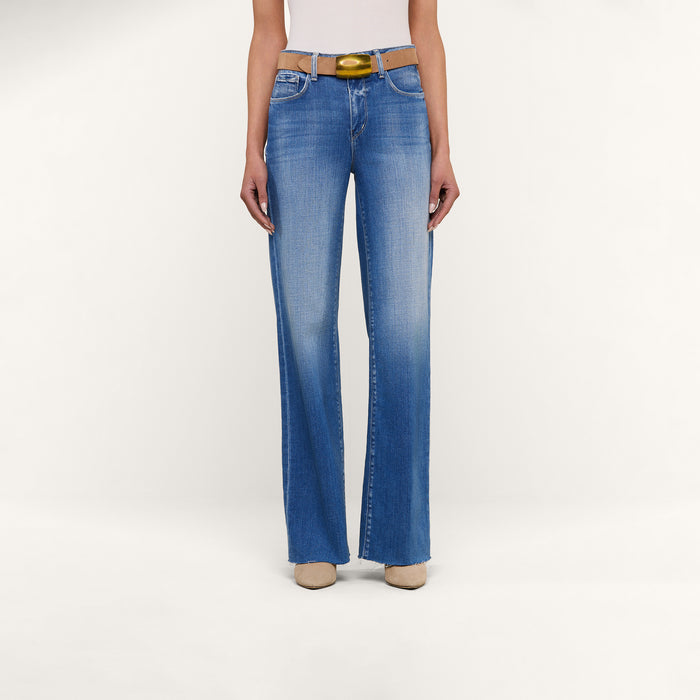 A person wearing the Scottie Wide-Leg Jean in light blue with a raw hem, paired with a white top, a yellow belt, and beige shoes stands against a plain white background.