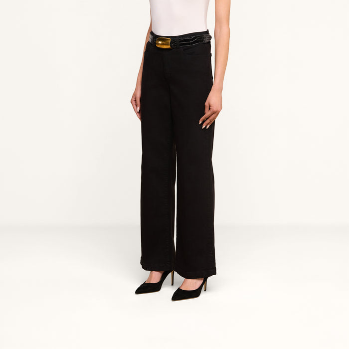A person stands against a plain white background wearing the Scottie Wide-Leg Jean in black high-rise premium stretch denim, paired with a white sleeveless top, black high heels, and a black belt with a gold buckle.