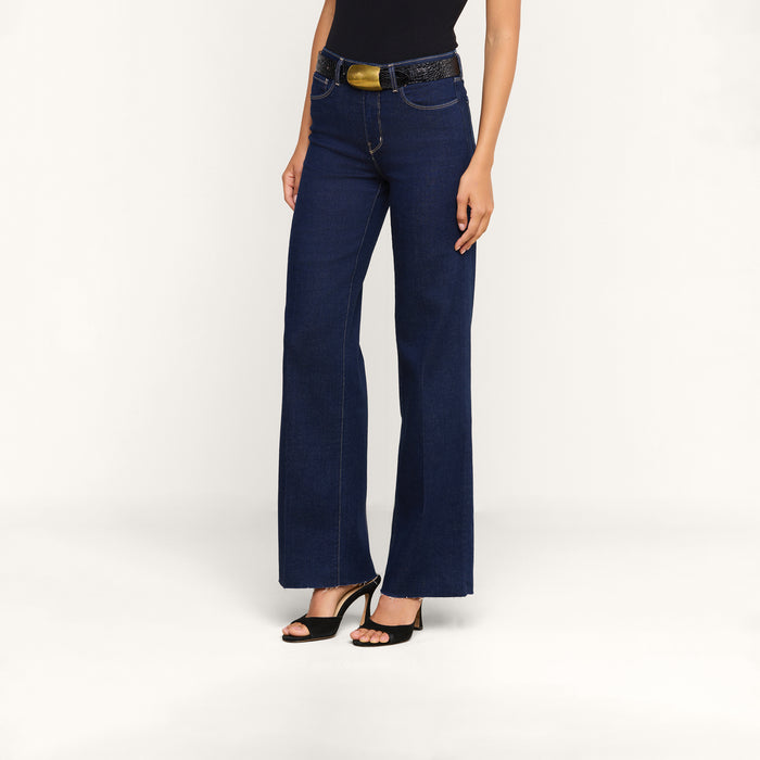 Wearing the Scottie Wide-Leg Jean in dark blue—crafted from premium stretch denim—a person stands against a plain white background, paired with a black belt, black sleeveless top, and black high-heeled sandals.