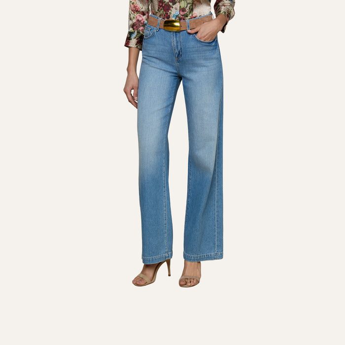 Wearing the Scottie Wide-Leg Jean—high-waisted with a light wash and contoured waistband—paired with a floral long-sleeve top and tan heeled sandals, this person stands against a plain background.