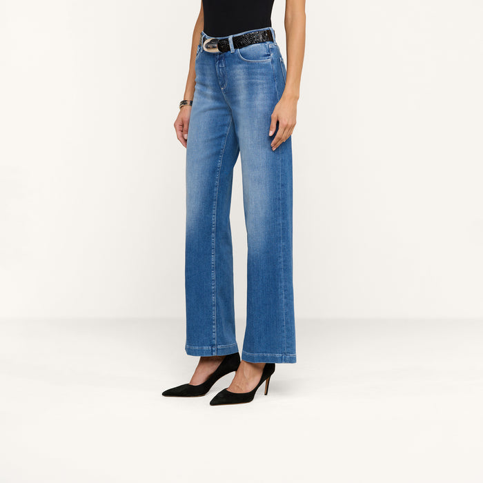 A woman is shown from neck to feet, standing against a plain white background, wearing the Scottie Wide-Leg Jean in blue stretch denim along with a black sleeveless top, a black belt, and black high heels.
