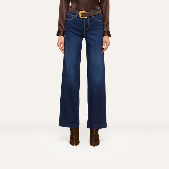 A person wearing high-waisted, wide-leg blue jeans and a brown long-sleeve blouse styled with a brown belt featuring a gold buckle and brown heeled boots stands confidently against a white background.