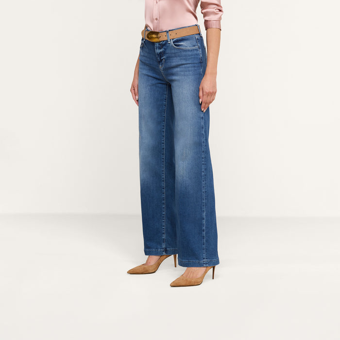 A person wearing a pink satin shirt, the Scottie Wide-Leg Jean in high-rise blue stretch denim, a brown belt, and tan high-heeled shoes stands against a plain white background.
