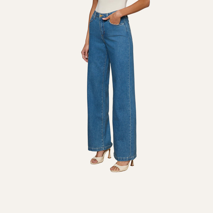 A person wearing the Scottie Wide-Leg Jean, a high-waisted, medium wash style made from 100 cotton denim, paired with open-toe beige heels stands against a plain, light background. Only the lower half of the body is visible.
