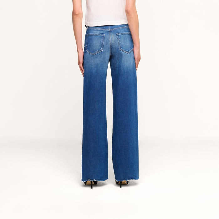 A person wearing a white top and the Scottie Wide-Leg Jean in vintage-wash blue denim, featuring high-rise, wide-leg silhouette with frayed hems and visible back pockets, stands with their back to the camera against a plain white background.
