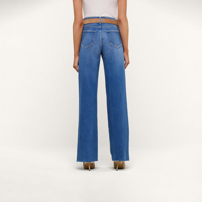 A person wearing a white top and the Scottie Wide-Leg Jean in a blue high-rise style stands facing away, showcasing the back pocket and seam details. The background is plain and light-colored.