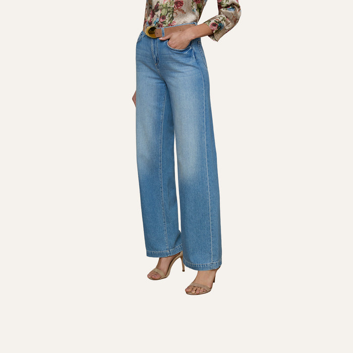 Wearing the Scottie Wide-Leg Jean, a contoured waistband blue jean, along with a floral long-sleeve blouse and heeled sandals, a person stands with one hand in their pocket against a plain light background.