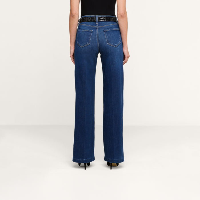 Wearing the Scottie Wide-Leg Jean in dark blue with a contoured waistband and back pockets, paired with a black sleeveless top, black belt, and black heeled shoes, a person stands facing away against a plain white background.