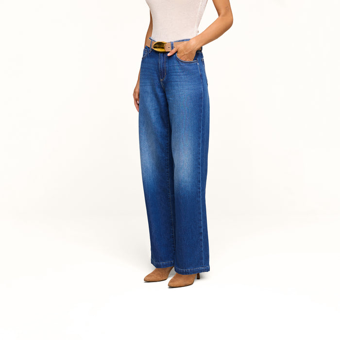 Woman wearing a sleeveless beige top, the Scottie Wide-Leg Jean in an indigo wash with a high-rise fit and black belt, paired with brown pointed-toe heeled shoes, standing against a plain white background.