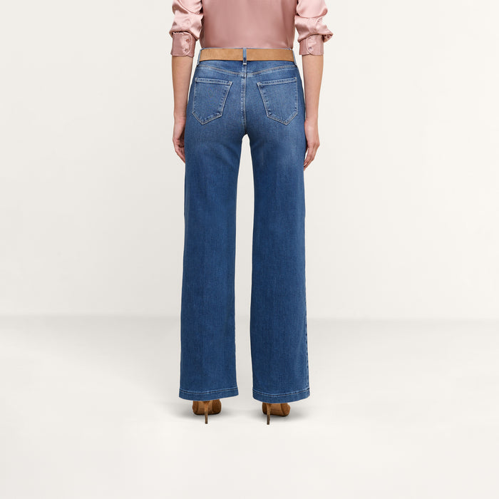 A person stands with their back to the camera, wearing a pink long-sleeve blouse, brown heeled shoes, and the Scottie Wide-Leg Jean in high-rise stretch denim, set against a plain light background.
