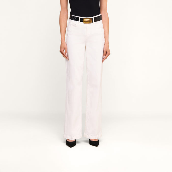 Wearing the Scottie Wide-Leg Jean in white, a person stands against a plain light background, styled with a black sleeveless top, a white belt with a gold buckle, and black pointed-toe shoes.