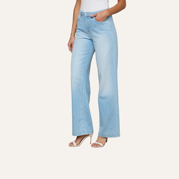 A person wearing the Scottie Wide-Leg Jean in light blue stretch denim, paired with a white sleeveless top and white high-heeled sandals, stands against a plain background.