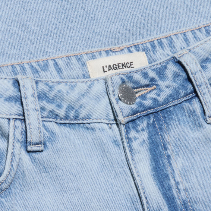 Close-up of the Scottie Wide-Leg Jean in a light wash, featuring a high-rise silhouette, visible silver button, and a white fabric label reading LAGENCE sewn into the waistband. The jeans are displayed against a blue denim background.