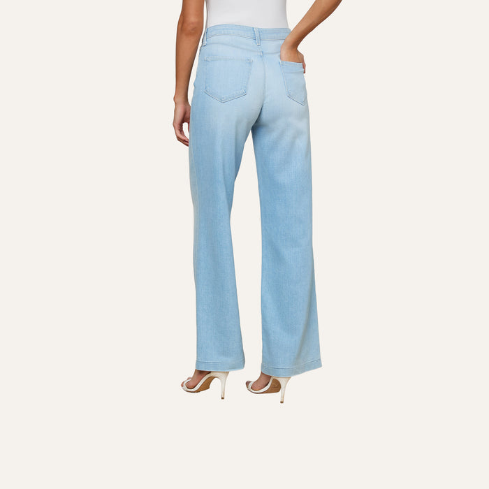 A woman is shown from the back wearing the Scottie Wide-Leg Jean in light blue stretch denim, paired with white high-heeled shoes; her left hand rests on her hip against a plain background.