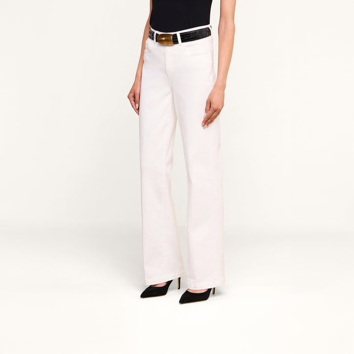 A person is wearing the Scottie Wide-Leg Jean, a high-waisted, wide-leg white pant with a high-rise waistband and a black belt, styled with a black sleeveless top and black high-heeled shoes, standing against a plain white background.