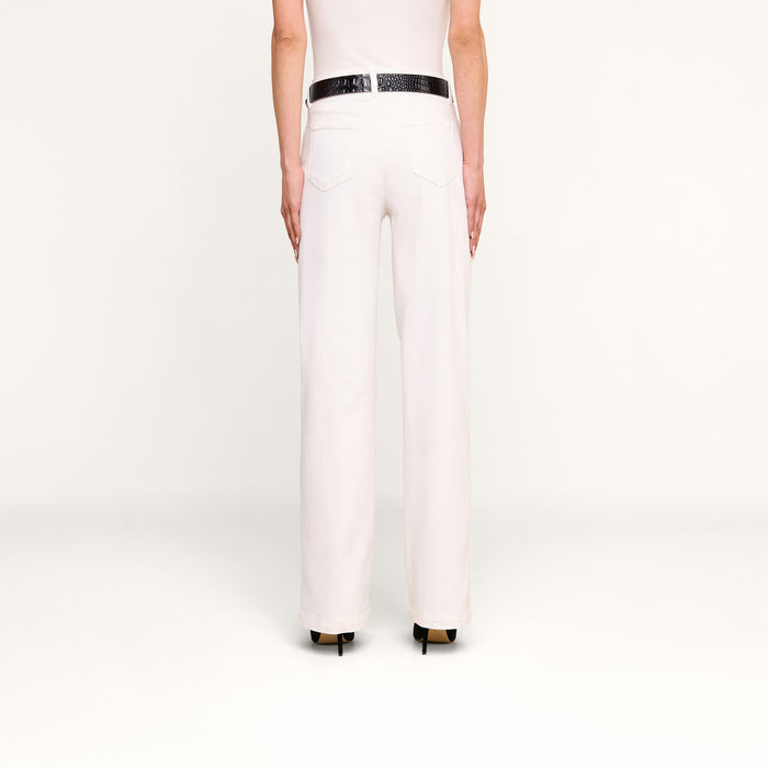 A person stands facing away against a plain white background, wearing the Scottie Wide-Leg Jean in white with a high-rise waistband, paired with a white top, a black belt, and black high-heeled shoes.