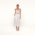 A woman stands against a white background wearing a white crop top and the Seina Poplin Midi Skirt—a high-waisted, ankle-length skirt featuring a small colorful print—paired with white high-heeled sandals.