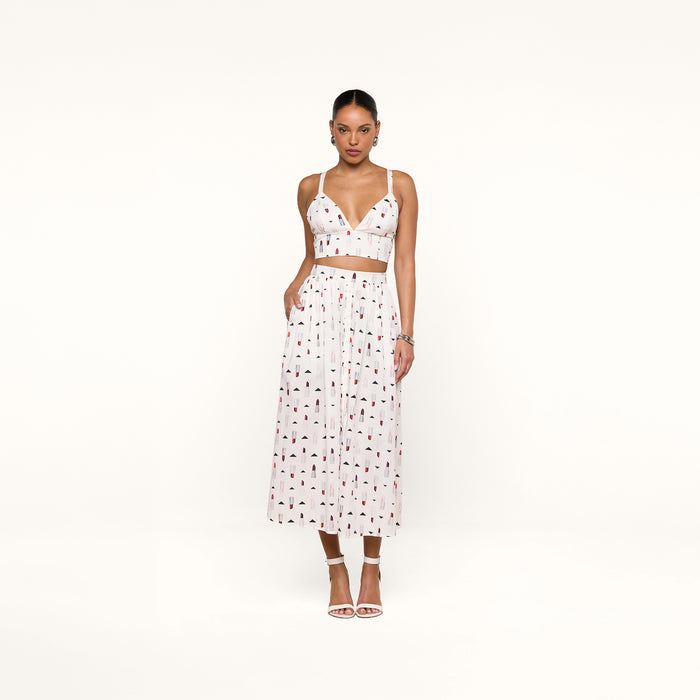 A woman stands against a white background wearing a white crop top and the Seina Poplin Midi Skirt—a high-waisted, ankle-length skirt featuring a small colorful print—paired with white high-heeled sandals.