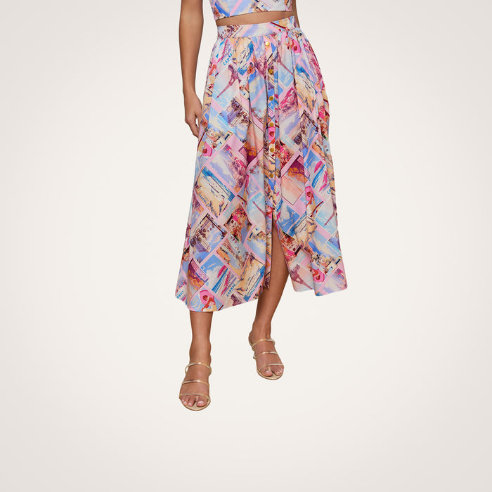 A woman models the Seina Poplin Midi Skirt, featuring a colorful geometric postcard print and a front slit, paired with strappy heeled sandals. Her upper body and face are not visible, and the background is plain white.