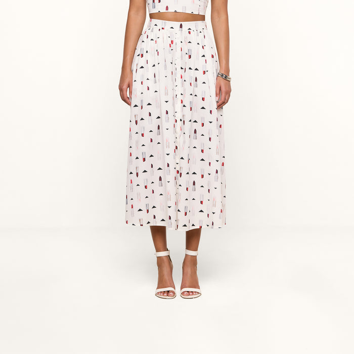 A woman stands against a plain white background wearing the Seina Poplin Midi Skirt, a high-waisted white skirt with a colorful geometric print, paired with a matching crop top, white ankle-strap heels, and a silver bracelet.