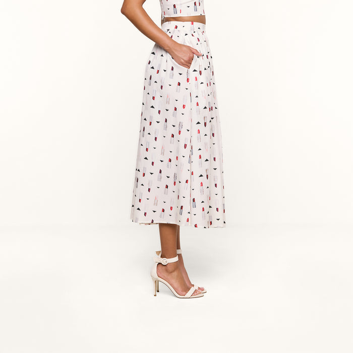 Sure Here’s a rewrite using the provided product name: A woman stands sideways against a white background, wearing the Seina Poplin Midi Skirt with a colorful lipstick print, paired with a matching crop top and blush high-heeled sandals, one hand tucked into her pocket.