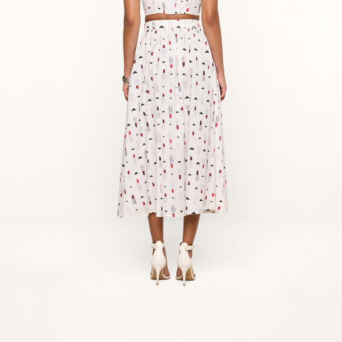 A woman stands facing away, wearing the Seina Poplin Midi Skirt—a white pleated skirt adorned with a colorful popsicle print—paired with white high-heeled sandals. The background is plain white.
