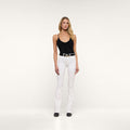 A woman with long blonde hair and a neutral expression stands against a plain white background, wearing the Selma Bootcut Jean with a contoured waistband, paired with a black sleeveless top, a black belt, and black shoes.
