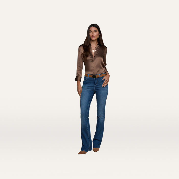 Woman with long hair wearing a brown satin blouse, the Selma Bootcut Jean in dark wash super stretch denim, a brown belt, and brown boots, standing against a plain white background.