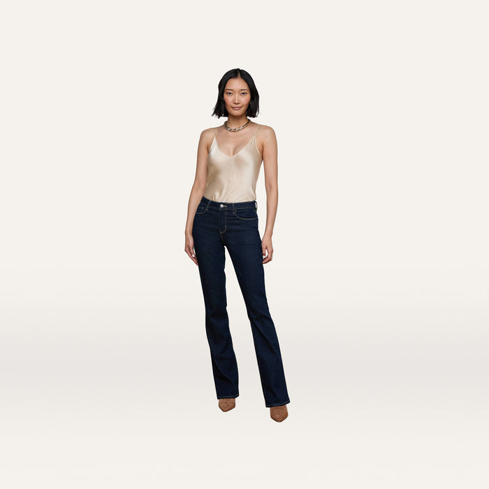 A person stands facing forward, wearing a beige satin camisole, the Selma Bootcut Jean—high-rise dark blue jeans made of stretch denim—and brown shoes, against a plain white background.